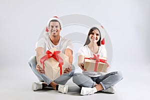 Beautiful happy couple in Santa hats sitting with Christmas gifts on light background