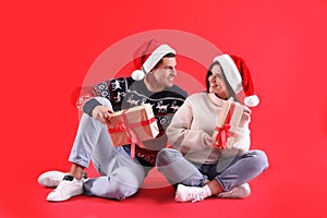 Beautiful happy couple in Santa hats and sweaters sitting with Christmas gifts on red background