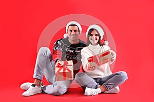 Beautiful happy couple in Santa hats and sweaters sitting with Christmas gifts on red background