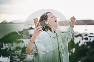 Beautiful happy curly-haired young girl hears music from smartphone in headphones and sings with city view