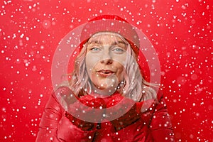 Beautiful happy girl blowing white snowflakes from her hands on red christmas background. Winter concept, winter fashion