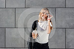 Beautiful happy girl with cold fresh smoothie outdoors. Smiling hipster girl with cup of milkshake on city street.