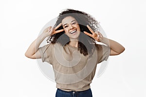 Beautiful happy girl enjoying life, showing peace v-sign and laughing carefree, standing upbeat against white background