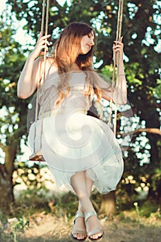 Beautiful happy girl with long hair swinging on rope swing on summer nature, young woman enjoy flying in foliage, leisure activity