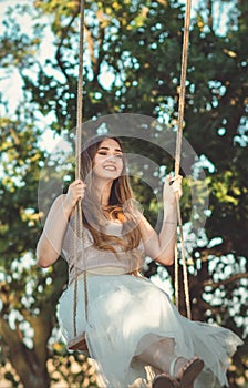 Beautiful happy girl with long hair swinging on rope swing on summer nature, young woman enjoy flying in foliage, leisure activity