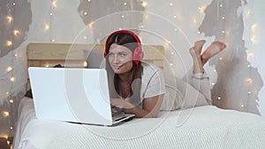 Beautiful happy girl is lying on a large bed using a white computer mobile computer and listening to music on headphones