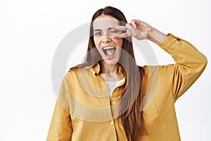 Beautiful happy girl shows peace v-sign over eye, winking and smiling broadly, express positive and joyful lifestyle