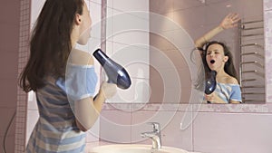 Beautiful happy girl teenager dries hair with hair dryer and sings and dances in front of a mirror in the bathroom