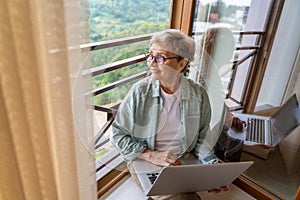 Beautiful happy gray haired senior mature woman in glasses using laptop while sitting by the window at home