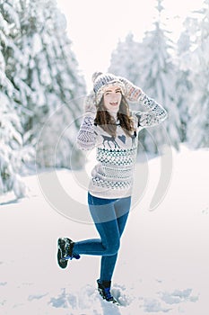 Beautiful happy laughing young woman wearing winter hat gloves and scarf covered with snow flakes. Winter forest landscape backgro