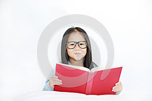 Beautiful happy little Asian kid girl reading hardcover book lying on bed against white background