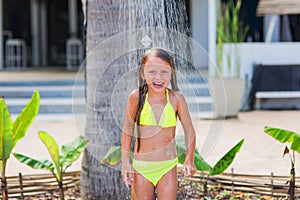 Beautiful happy little girl in a yellow bathing suit under a shower on the beach in a tropical garden