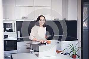 Beautiful happy mature elderly woman with orange in her hands standing at home in the kitchen in front of laptop screen, healthy