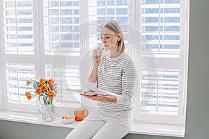 Beautiful happy middle age woman sitting on windowsill at home and reading book. Young woman with short hair eating macaroons and