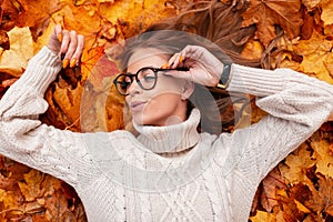 Beautiful happy model of a young woman in a stylish sweater in a trendy glasses lies and enjoys rest on the orange autumn leaves