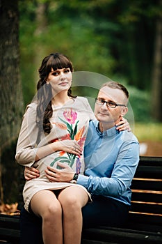 Beautiful and happy pregnant couple relaxing outside in the autumn park sitting on bench.