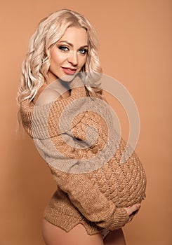 Beautiful happy pregnant woman expecting maternity hugging her belly. Blonde in knit sweater isolated on studio beige background.