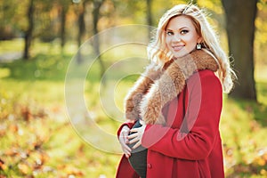 Beautiful happy pregnant woman staying in autumn park touching her belly and enjoying expecting of baby.