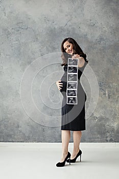 Beautiful happy pregnant woman with ultrasound scan image standing on gray background