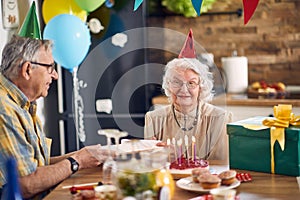 Beautiful happy senior couple sitting at kitchen table in birthday hats, elderly man giving a birthday present to her wife