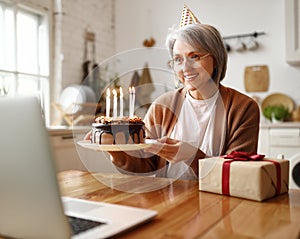 Beautiful happy senior woman with cake with lit candles , celebrating birthday online with family