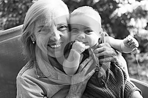 Beautiful happy smiling senior elderly woman holding on hands cute little baby boy sitting outdoor. Grandmother grandson