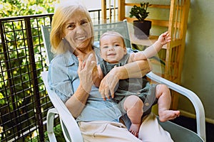 Beautiful happy smiling senior elderly woman holding on hands cute little baby boy sitting on outdoor rocking chair