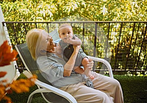 Beautiful happy smiling senior elderly woman holding on hands cute little baby boy sitting on outdoor rocking chair