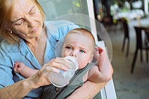 Beautiful happy smiling senior elderly woman nanny feeding formula on hands from bottle cute little baby boy