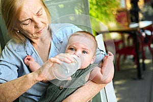 Beautiful happy smiling senior elderly woman nanny feeding formula on hands from bottle cute little baby boy