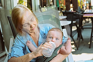 Beautiful happy smiling senior elderly woman nanny feeding formula on hands from bottle cute little baby boy