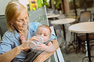 Beautiful happy smiling senior elderly woman nanny feeding formula on hands from bottle cute little baby boy