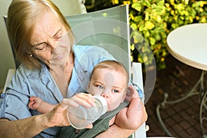Beautiful happy smiling senior elderly woman nanny feeding formula on hands from bottle cute little baby boy