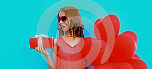 beautiful happy smiling woman holding gift box and bunch of red heart shaped balloons on a blue background