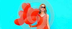 Beautiful happy smiling young woman with bunch of red heart shaped balloons wearing sunglasses on blue background