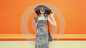 Beautiful happy smiling young woman with cup of coffee wearing black summer straw hat, striped dress with handbag on orange