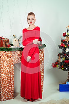 Beautiful happy smiling young woman in evening dress with bright makeup with red lipstick sitting near the Christmas tree
