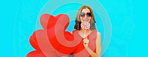 Beautiful happy smiling young woman with lollipop and bunch of red heart shaped balloons wearing sunglasses on blue background