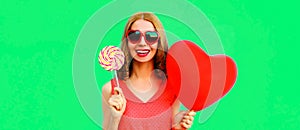 Beautiful happy smiling young woman with lollipop and bunch of red heart shaped balloons wearing sunglasses on green background