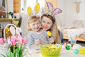 Beautiful happy son and mother in headbands with bunny ears decorate eggs with a brush and bright paints, prepare to