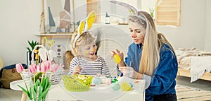 Beautiful happy son and mother in headbands with bunny ears decorate eggs with a brush and bright paints, prepare to