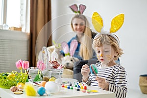 Beautiful happy son and mother in headbands with bunny ears decorate eggs with a brush and bright paints, prepare to