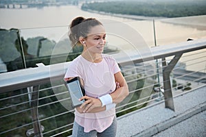 Beautiful happy sporty girl with earphones standing with crossed arms on the city bridge and smiling toothy smile looking away.