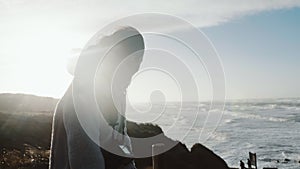 Beautiful happy tourist woman with camera standing under extreme cold wind covering face at winter sea shore slow motion