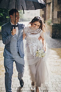 Beautiful happy wedding couple embracing together under umbrella in sunny rainy street. Stylish bride and groom walking and