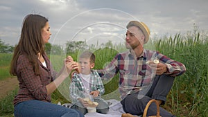 Beautiful happy woman feeds man while relaxing with child at family picnic on open air in green field close up