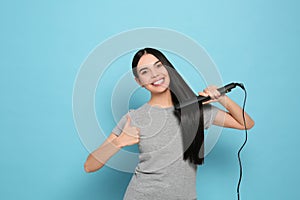 Beautiful happy woman showing thumbs up while using hair iron on light blue background. Space for text