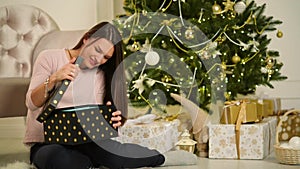 Beautiful Happy Woman Sitting Next To Christmas Tree With A Gift In Her Hands. Merry Christmas and New Year Concept.