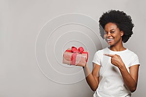 Beautiful happy woman smiling, holding gift box and pointing at red present on white background