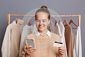 Beautiful happy woman wearing beige sweater, holding with credit card and cell phone, entering data and paying for shopping online